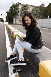 Smiling young woman with skateboard sitting on bollard using smartphone - GIOF07688