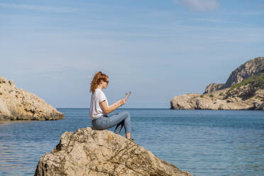 Rothaarige Frau sitzt auf einem Felsen und liest eine Zeitschrift - AFVF04194