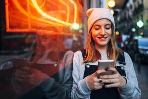 Lächelnde junge Frau, die neben einem Neonlicht steht und ihr Smartphone bei Nacht benutzt - JCMF00283