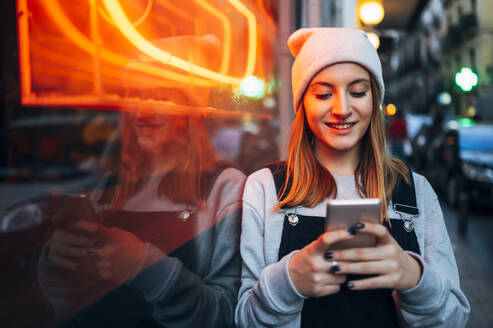 Smiling young woman standing next to neon light using her smartphone at night - JCMF00283