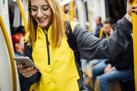 Lächelnde junge Frau steht in der U-Bahn und benutzt ihr Smartphone, lizenzfreies Stockfoto