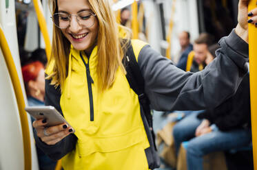 Lächelnde junge Frau steht in der U-Bahn und benutzt ihr Smartphone - JCMF00291
