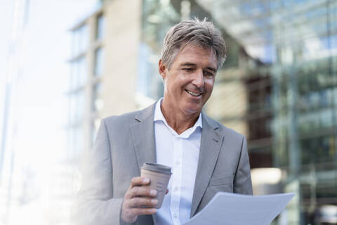 Lächelnder Geschäftsmann mit Kaffee zum Mitnehmen bei der Durchsicht von Dokumenten in der Stadt, lizenzfreies Stockfoto