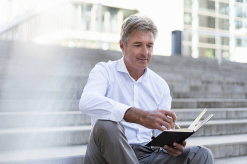 Mature businessman sitting on stairs looking at notebook - DIGF08949