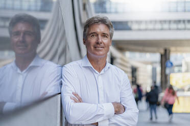 Portrait of confident mature businessman leaning against a glass front in the city - DIGF08955