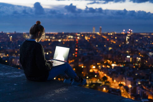 Junge Frau mit Laptop in der Morgendämmerung über der Stadt, Barcelona, Spanien - GIOF07691