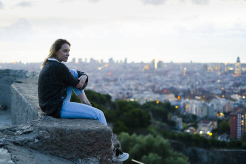 Junge Frau sitzt bei Sonnenaufgang über der Stadt, Barcelona, Spanien - GIOF07697