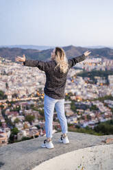 Rear view of carefree young woman standing above the city at sunrise, Barcelona, Spain - GIOF07698