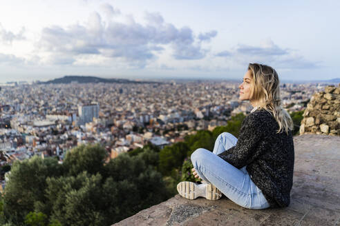 Junge Frau sitzt bei Sonnenaufgang über der Stadt, Barcelona, Spanien - GIOF07705