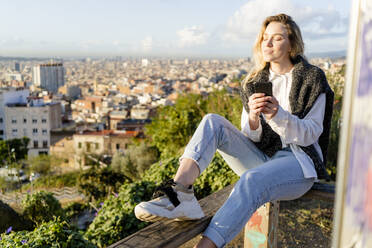 Junge Frau sitzt auf einem Geländer über der Stadt und hält ein Mobiltelefon in der Hand, Barcelona, Spanien - GIOF07718
