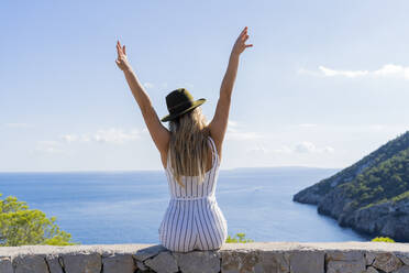 Junge Frau, die mit erhobenen Armen auf einer Mauer sitzt und in die Ferne schaut, Aussichtspunkt in Ibiza - AFVF04224