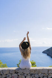Junge Frau, die mit erhobenen Armen auf einer Mauer sitzt und in die Ferne schaut, Aussichtspunkt in Ibiza - AFVF04225