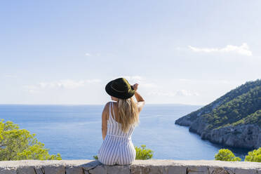 Junge Frau auf einer Mauer sitzend, die Hand auf dem Hut, Aussichtspunkt in Ibiza - AFVF04227