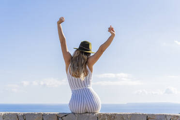 Junge Frau, die mit erhobenen Armen auf einer Mauer sitzt und in die Ferne schaut, Aussichtspunkt in Ibiza - AFVF04228