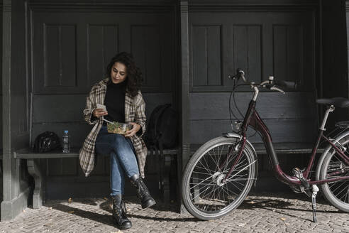 Frau mit Fahrrad beim Mittagessen und mit Smartphone auf dem Bahnsteig, Berlin, Deutschland - AHSF01203