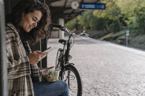 Lächelnde Frau mit Fahrrad beim Mittagessen und mit Smartphone auf dem Bahnsteig, Berlin, Deutschland - AHSF01204