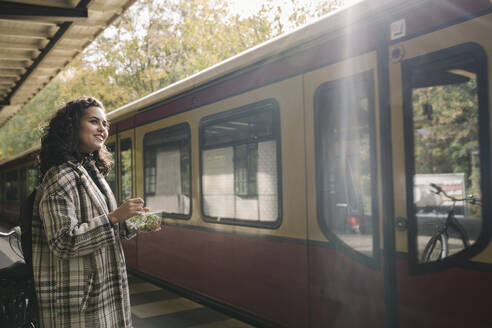 Lächelnde Frau beim Mittagessen auf dem Bahnsteig einer U-Bahn-Station, Berlin, Deutschland - AHSF01209