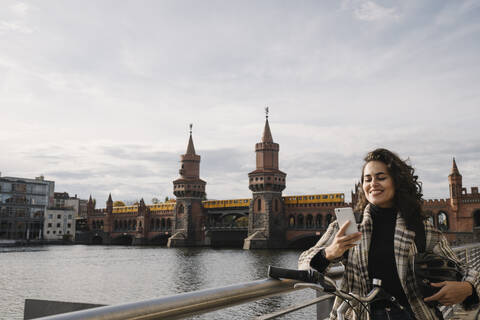 Lächelnde Frau mit Fahrrad und Smartphone in der Stadt an der Oberbaumbrücke, Berlin, Deutschland, lizenzfreies Stockfoto