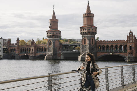 Lächelnde Frau mit Fahrrad und Smartphone in der Stadt an der Oberbaumbrücke, Berlin, Deutschland, lizenzfreies Stockfoto