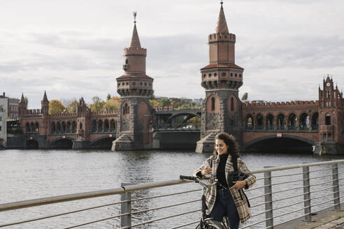 Lächelnde Frau mit Fahrrad und Smartphone in der Stadt an der Oberbaumbrücke, Berlin, Deutschland - AHSF01233