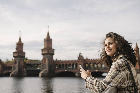 Lächelnde Frau mit Smartphone in der Stadt an der Oberbaumbrücke, Berlin, Deutschland - AHSF01235