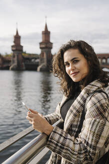 Porträt einer Frau mit Smartphone in der Stadt an der Oberbaumbrücke, Berlin, Deutschland - AHSF01236