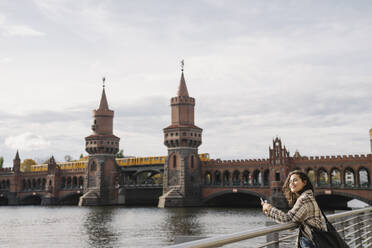 Lächelnde Frau mit Smartphone in der Stadt an der Oberbaumbrücke, Berlin, Deutschland - AHSF01237