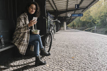 Woman with bicycle having lunch and using smartphone on station platform, Berlin, Germany - AHSF01242