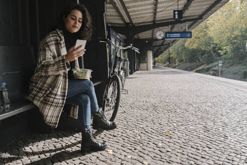 Frau mit Fahrrad beim Mittagessen und mit Smartphone auf dem Bahnsteig, Berlin, Deutschland - AHSF01242