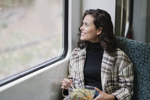 Lächelnde junge Frau beim Mittagessen in einer U-Bahn mit Blick aus dem Fenster - AHSF01246