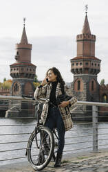 Smiling woman with bicycle on the phone in the city at Oberbaum Bridge, Berlin, Germany - AHSF01256