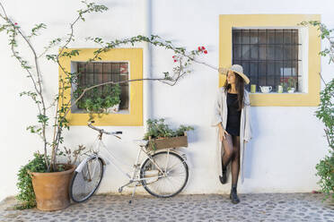 Junge Frau mit Sonnenhut und Strickjacke lehnt an einer Wand - AFVF04244