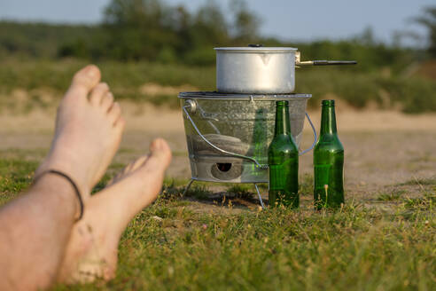 Nackte Füße eines jungen Mannes vor einem Campingkocher und leeren Bierflaschen - LBF02792