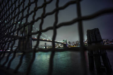 USA, New York, New York City, Maschendrahtzaun gegen beleuchtete Brooklyn Bridge bei Nacht - OCMF00915