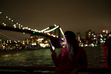 USA, New York, New York City, Junge Frau mit Smartphone vor beleuchteter Brooklyn Bridge bei Nacht - OCMF00917
