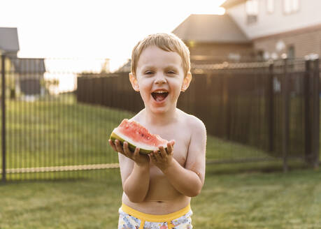 Porträt eines fröhlichen Jungen, der im Hinterhof eine Wassermelone isst - CAVF69248