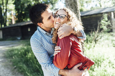 Man kissing cheerful girlfriend while standing in backyard - CAVF69352