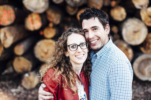 Porträt eines glücklichen Paares, das vor Holzstämmen steht - CAVF69353