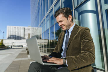 Happy businessman sitting in urban business district using laptop, Madrid, Spain - KIJF02732