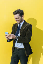 Glücklicher Geschäftsmann mit Smartphone vor gelber Wand - KIJF02744