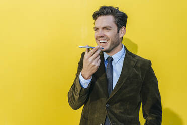 Happy businessman using smartphone in front of yellow wall - KIJF02745
