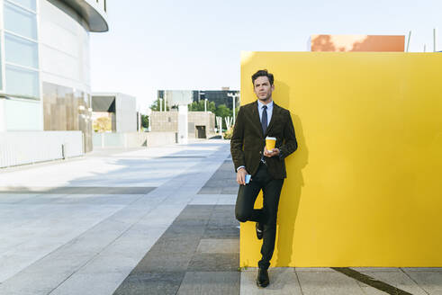 Businessman with takeaway coffee and phone next to yellow wall in urban business district, Madrid, Spain - KIJF02746