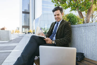 Businessman sitting on bench in the city writing in notebook and looking at laptop, Madrid, Spain - KIJF02757