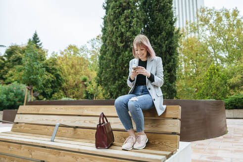 Lächelnde Frau sitzt auf einer Parkbank und benutzt ein Mobiltelefon - KIJF02795