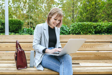 Lächelnde Frau sitzt auf einer Parkbank und benutzt einen Laptop - KIJF02797