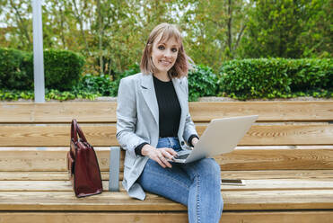 Lächelnde Frau sitzt auf einer Parkbank und benutzt einen Laptop - KIJF02798