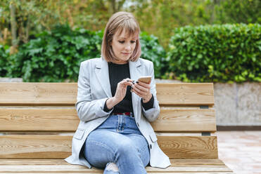 Frau sitzt auf einer Parkbank und benutzt ein Mobiltelefon - KIJF02799