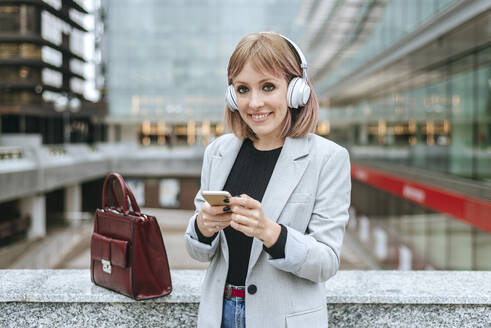 Lächelnde Frau mit Smartphone und Kopfhörern in der Stadt - KIJF02803