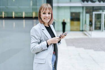 Porträt einer lächelnden Frau mit Mobiltelefon an einem Glasgebäude - KIJF02817
