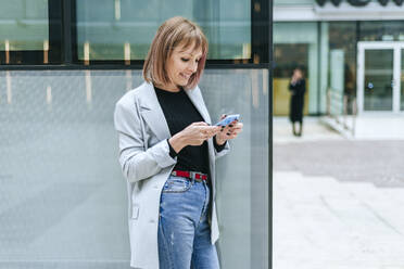 Lächelnde Frau, die ein Mobiltelefon an einem Glasgebäude benutzt - KIJF02819